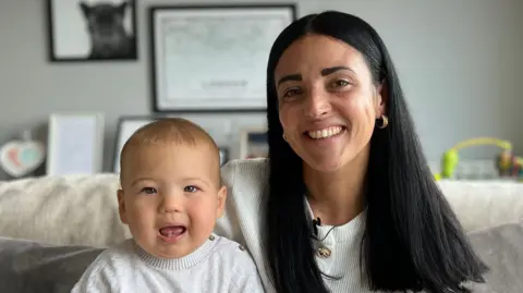 Lauren is sitting on a sofa holding a Nico on her lap, both facing the camera. Framed pictures hang on the wall behind them, and soft furnishings and baby toys are visible in the living room.
