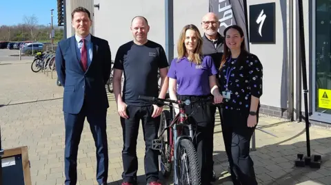 Blaby District Council People stood gathered around a bike in front of a building. Three are three men and two women pictured. 

