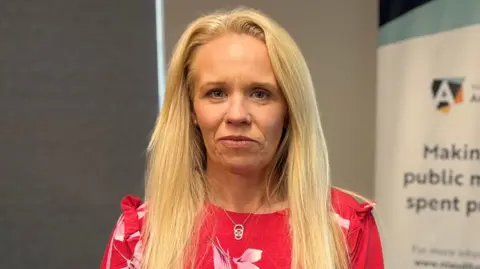 Dorinnia looking at the camera. She is wearing a red top with pink flowers on it. She has long blonde hair.