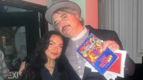 Georgia Griffiths Georgia stands and smiles next to Pete who is holding a copy of her poetry book. Pete, on the right, wears a striped shirt, black suit jacket and a grey hat. He has his arm over Georgia, who wears a denim jumpsuit and has curly long black hair.
