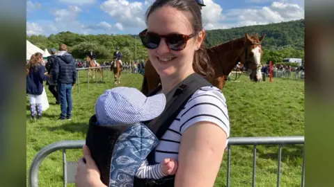 HANDOUT Gina, who has brown long hair in a ponytail and its wearing tortoiseshell sunglasses and a white and black striped t-shirt, pictured holding her baby son in a baby carrier