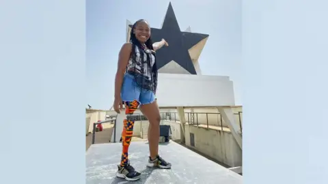 Abena Christine Jon'el standing on a platform with a prosthetic right leg wrapped in kente cloth. She is pointing behind her at the top of Accra's Black Star monument.