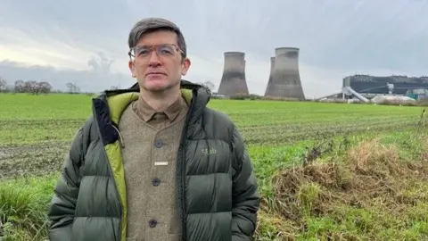 Professor Richard Brook has short hair and is wearing a green jacket over a tweed jacket and is standing in front of the Fiddler's Ferry cooling towers