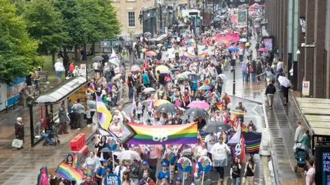 Pride march - hundreds of people wearing bright colours, waving rainbow flags  - a symbol of lesbian, gay, bisexual, transgender and queer pride - and carrying banners marching down a street. The weather is raining and some are sporting umbrellas. People are standing on the pavement watching them.