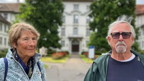 Lola and David are standing outdoors in front of a large, white building with a red roof and multiple windows. The building is surrounded by trees and greenery. The person on the left is wearing a light blue patterned jacket and has short hair, while the person on the right is wearing a dark blue shirt with a green jacket over it. The sky appears to be cloudy.