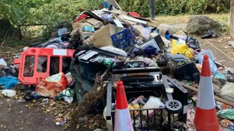 A large pile of waste sits at the back of a laneway. There are bollards and other street construction gear in among the rubbish.