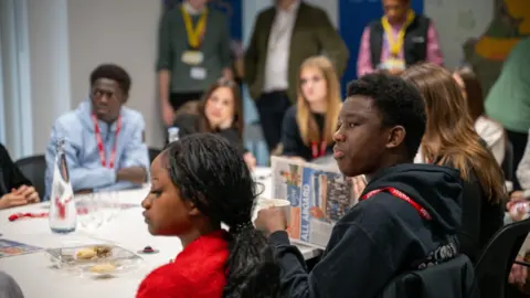 ISCRE A group of young people sit around a table in discussion. One is reading a newspaper and others are looking to the front of the table. 