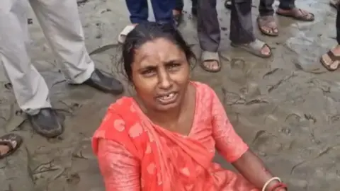 A woman wearing an orange saree sitting on wet sand with the feet of people standing behind in the frame 