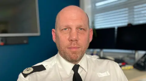 Head and shoulders photo of ACC Tony Rowlinson who is bald, bearded and wears a police shirt and tie.