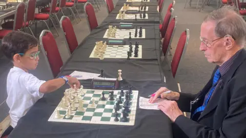 A young boy wearing a white shirt and colourful glasses, with short dark hair sits on a red chair at a table opposite a man with grey hair, wearing a suit and glasses, with a newspaper rolled up in his pocket. They are playing chess. There are many other chess boards and chairs lined up along tables in the room.