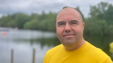 A man in a bright yellow T-shirt stands in front of a lake with green trees in the background under a grey sky. He has cropped dark hair and light stubble. 