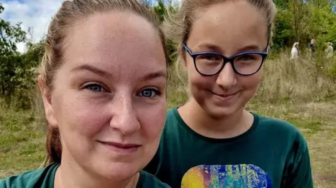 Kim/BBC Close-up of a white mum and daughter's faces as they stand in a field. The daughter is wearing glasses with blue frames. Three figures stand in the distance under a tree.