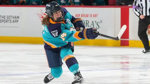 Jaime Bourbonnais lifts her hockey stick in what appears to be the aftermath of a shot. She is wearing a teal, navy and orange uniform and her ponytail is sticking out behind her helmet. In the back right corner of the image, a referee is visible 
