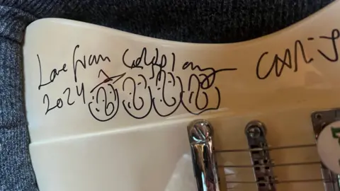 A white electric guitar is signed by Coldplay in black marker. It says Love from Coldplay 2024. There is a drawing of four faces next to it.