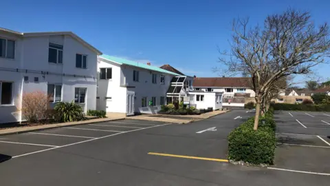 BBC Raymond Falla House - A building with a green roof, snaking down the left of the photo. It is painted white. There is a car park with a row of trees on the right of the image. 
