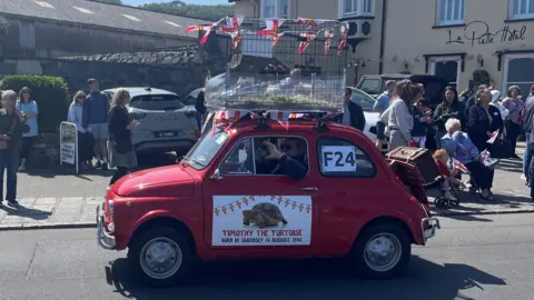 A red classic fiat 500 taking part in the cavalcade. A large cage is on top of the car. Inside you can see the tortoises shell and some staw.