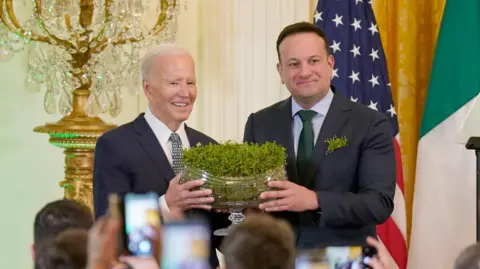 Pa Media Leo Varadkar stands on the stage with a large crystal bowl from Shamrocks. He delivers it to Joe Biden, who stands on his right. They both smile because people take pictures.