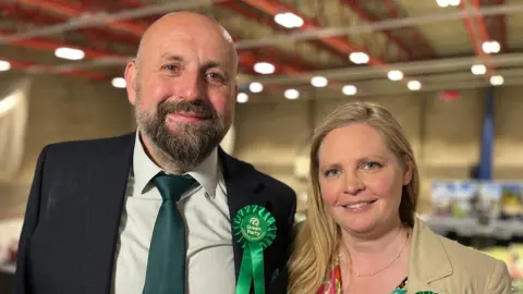 Stephen Ferguson and Lara Davenport-Ray are standing side by side. Ferguson, who is on the left, has a short brown and grey beard. He is wearing a cream shirt, green tie and black suit jacket. He is wearing a Green Party rosette. Davenport-Ray is on the right. She has long blonde hair and is wearing a floral dress and beige blazer.