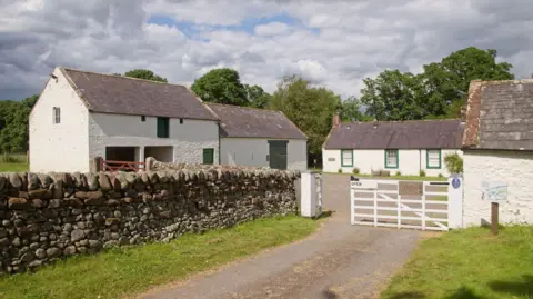 Ian Findlay White farm buildings surrounded by a stone wall. Trees are in the background and there is a white fence and gate at the entrance.