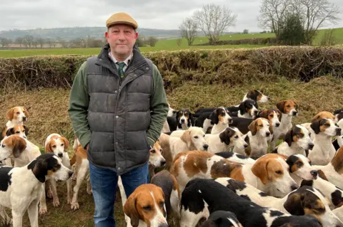 Tim Hardy is standing in a hedgerow in the Cumbrian countryside surrounded by foxhounds. He is wearing a flat cap, a shirt and tie and a body warmer.