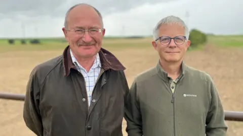 Nick Acklam and Tony Day in front of a field. Nick has glasses and a moustache and is wearing a dark green coat and white shirt with blue check. Tony has short white hair, rounded glasses and is wearing a dark green fleece.