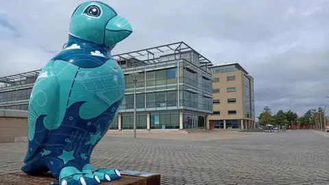 Puffins Galore One of the sculptures in the Puffins Galore trail. It is blue, green and white and standing on a low plinth. You can see modern office buildings behind it and it is situated on a pedestrianised walkway.