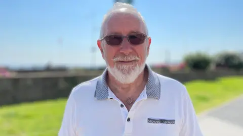Bill Holgate has short white hair and a beard. He's wearing sunglasses and a white polo shirt, in front of a strip of grass on a sunny day.