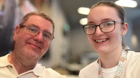 Jarman family Shane and Emma seated close together indoors, facing the camera in what appears to be a casual selfie or close-range photograph. Shane on the left is wearing a light-coloured polo shirt and glasses. He is seated slightly closer to the camera and occupies the left side of the frame. Emma on the right is wearing glasses and a white top. Around her neck is a patterned lanyard decorated with pastel-coloured flowers and cupcake illustrations, along with a delicate necklace and small star-shaped earrings.
