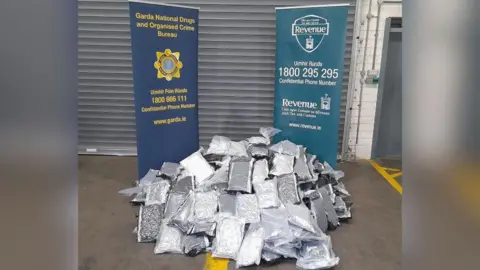 Multiple silver parcels are piled up on the ground in front of two blue signs, one for the Garda National Drugs and Organised Crime Bureau and the other for Irish Revenue. 