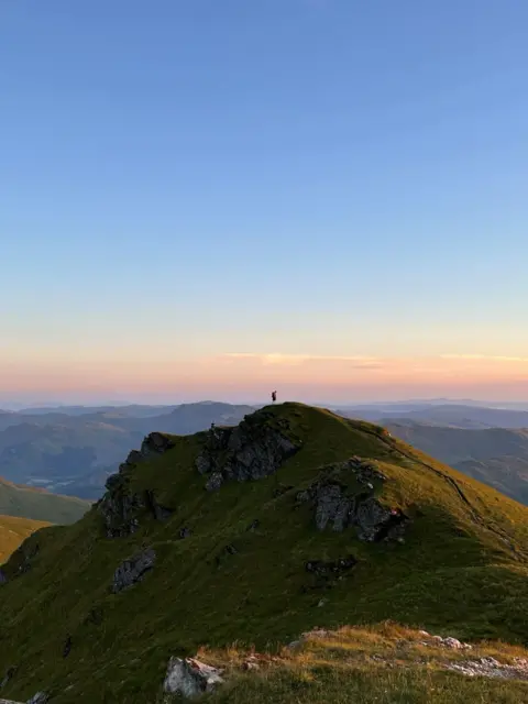 Dylan Weir A mountain with hills in the background. A person is on top of the mountain