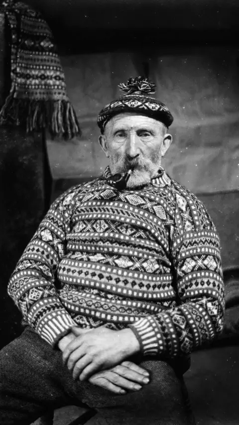 Shetland Museum & Archives An elderly man posing for a portrait. He is wearing a fair isle jumper and hat, and is smoking a pipe. 