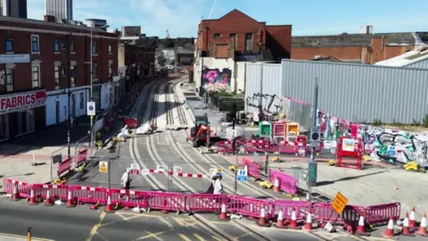 An empty street, viewed from the air, with red brick buildings down either side and pink barriers and traffic cones blocking it off at the near end