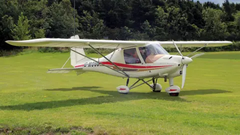 Derek Heley A small plane parked on grass. Two people can be seen outlined in the cockpit. There are trees behind the plane and it is a sunny day. The plane is small, white with some red stripes on it. It has "G-MOOD" written on its side. 