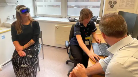 BBC A woman sits on a chair with a virtual reality headset on whilst a doctor and nurse look at a laptop screen.