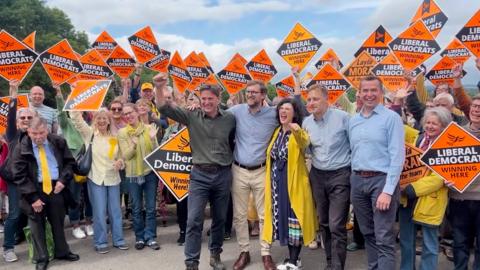 A Liberal Democrat photo op in Botley, with Layla Moran, Charlie Maynard, Calum Miller, Olly Glover, and Freddie van Mierlo celebrating in front of supporters holding Lib Dem placards
