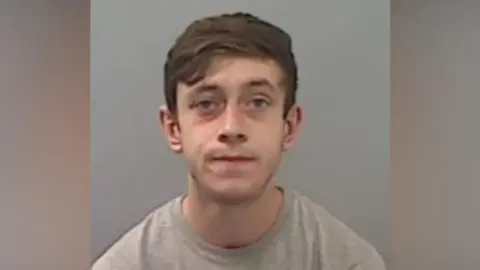 Cleveland Police Police mugshot of Harrison Simpson. He is a young man with short brown hair swept across his forehead and is slightly grimacing, wearing a grey t-shirt.