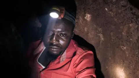 AFP A man in a red overall in an underground mine looks downward as a torch on his helmet illuminates what's in front of him.