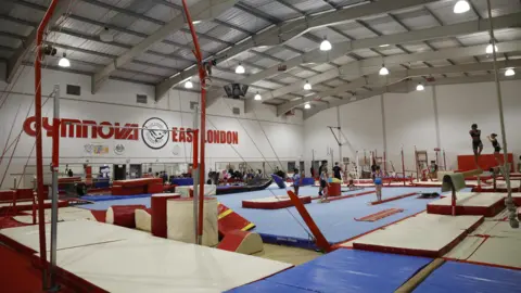 Facundo Arrizabalaga The inside of East London Gymnastic Centre. It is a large hall with gym equipment and mats filling the space. There are gymnasts on the equipment. In the background are the words "GYMNOVA EAST LONDON" in large red lettering along a white-painted wall.