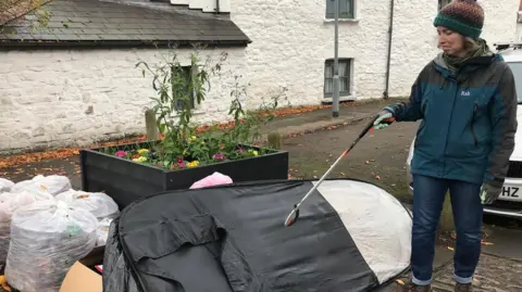 Fiona McAllister A spray tan booth and lots of full white bin bags in a pile beside a flower bed with a woman holding a litter picker to the right. She is wearing a bobble hat and waterproof jacket and has a look of disgust on her face as she looks at the fly-tipped item