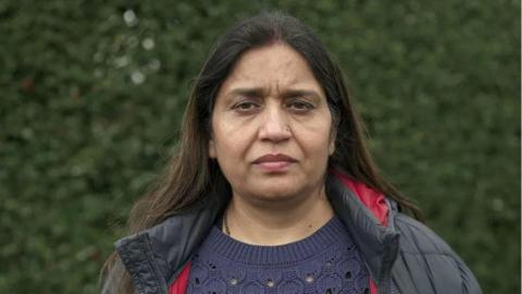 Seema Misra looks directly at the camera, she is wearing a black padded jacket, navy blue jumper and is standing in front of a hedge