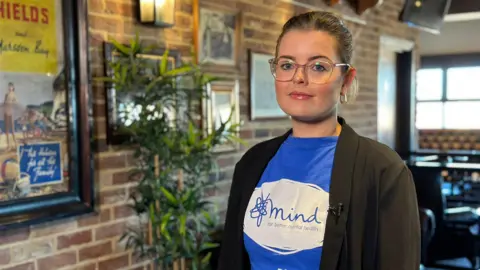 A photo Stephanie Cochrane who has brown hair tied back and clear framed glasses. She's wearing a Mind charity t-shirt and a black blazer.