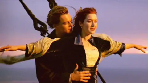 CBS via Getty Images A screen grab from the Titanic film. A man and woman stand at the bow of a ship with their arms outstretched against a dusky sky in an iconic scene from the 1997 film Titanic. The woman is in the foreground wearing a dark jacket, while the man stands behind her with his arms mirroring hers. Rigging and chains are visible to the left of the frame, with a calm ocean stretching to the horizon.