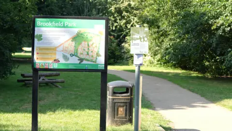The entrance to a park, with a map displayed on a board and a black bin to the left of the entrance path
