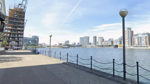 Waterfront walkway beside Royal Victoria Dock, with railings, lamp posts, modern flats and a building under construction on a clear day.