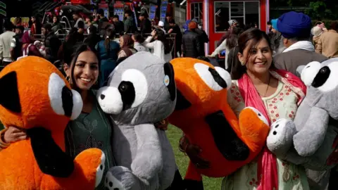 Two women smile at the camera. They hold two massive cuddly toys in each arm - one is a red panda and the other is a raccoon.