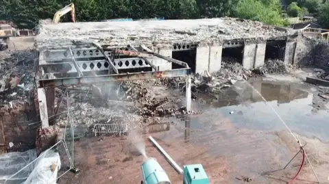 Anglian Demolition Water spraying at the derelict factory which was on fire and now is being demolished. It is a large building with the walls missing and steal work exposed. there are piles of broken concreat next to it. The picture is taken from above.