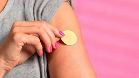 Getty Images A woman places a nicotine patch on her left upper arm. She has painted, pink nails and wears a grey t-shirt. The background is pink.