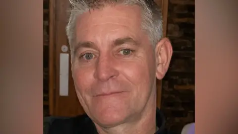 Close-up headshot of a man with short grey hair, looking toward the camera. The image is tightly cropped around the face, with a wooden interior background visible behind. The person is wearing a dark top and is photographed indoors under warm lighting.