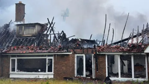 The burnt out remnants of a bungalow. Its roof has been destroyed with only a few frayed wooden beams remaining and set against thick grey smoke rising from the property. Many of the windows of the bungalow have shattered as a result of the intense heat from the fire. A lone firefighter on an aerial appliance is faintly visible on the other side of the property, through the smoke.