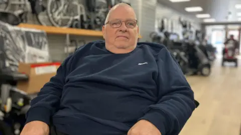A man in a wheelchair looks at the camera. In the background is the newly reopened Easy Mobility Services showroom. There are mobility scooters and other mobility support vehicles around him.